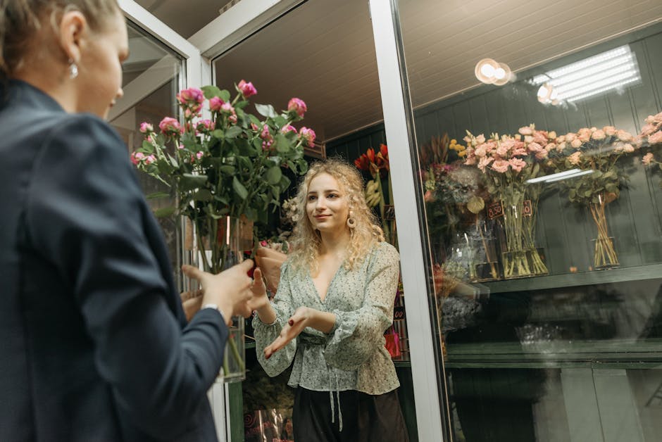A florist at flowerswatford.org.uk is handing a fresh bouquet of pink roses with green foliage, wrapped in brown paper, to a customer through a glass window at the shop. The customer, a young woman wi