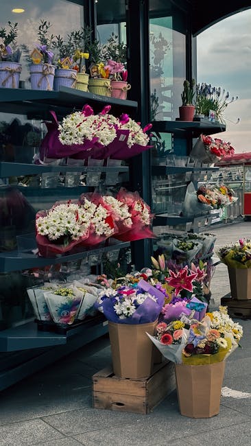 A display of vibrant floral arrangements and bouquets arranged on metal shelves and standing on the ground outside a flower shop. The flowers include white daisies, pink lilies, purple irises, and mix
