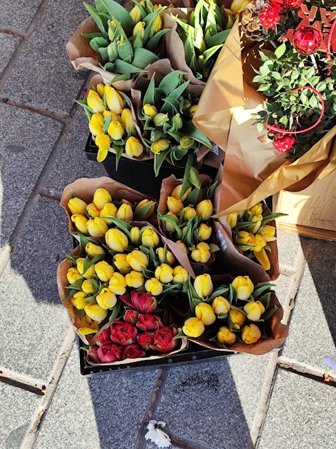 A high-angle view of three bouquets of fresh flowers arranged outdoors on a paved surface. The first bouquet contains yellow tulips with green stems and leaves, wrapped in brown paper. The second bouq